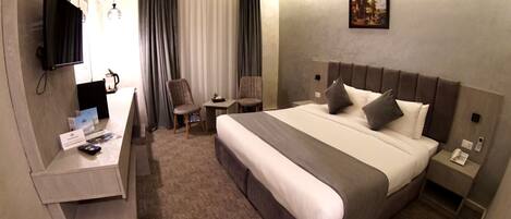 A hotel room with a large bed, a desk with a flat-screen TV, two chairs, a small table, and a painting on the wall.