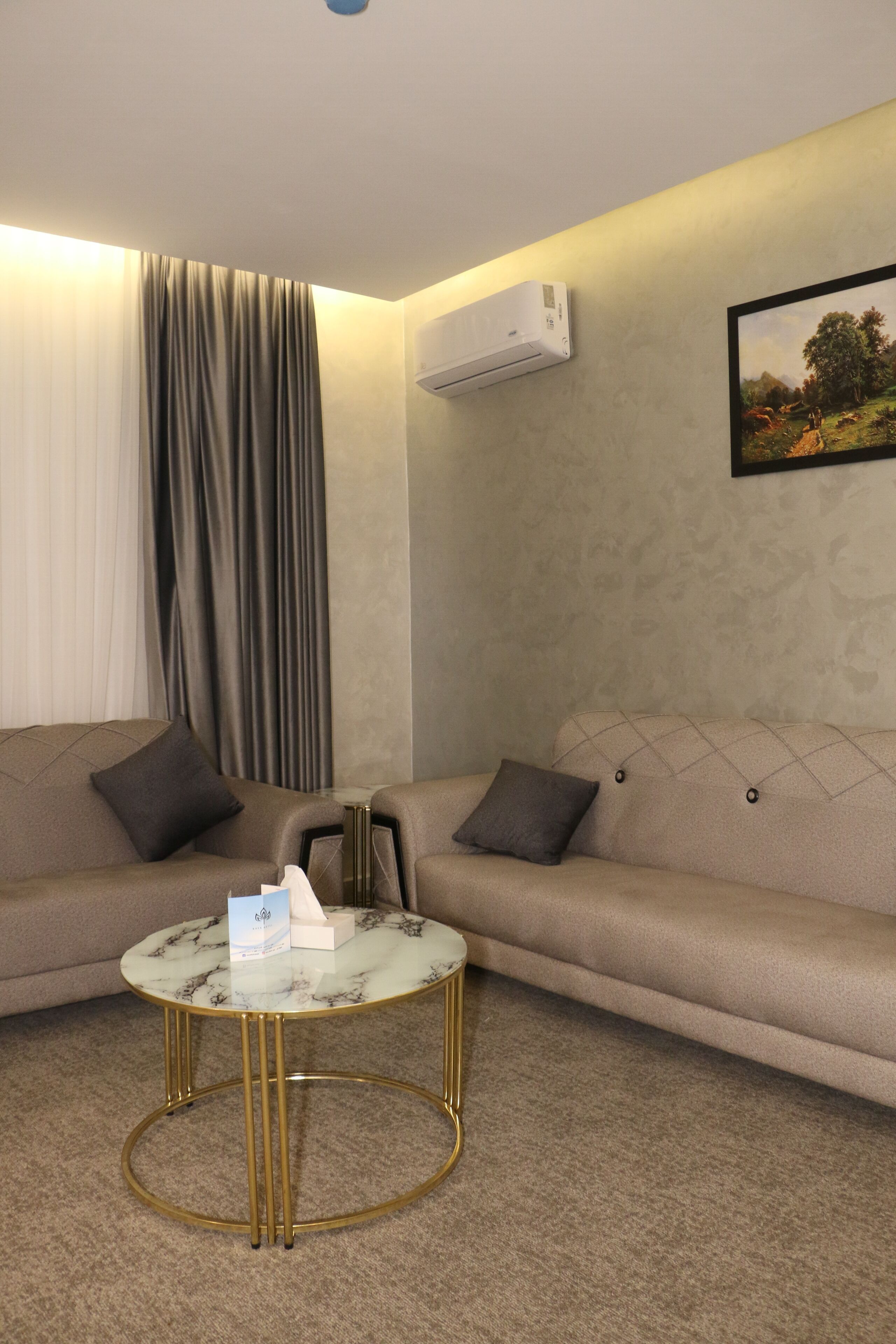 Ein modernes Wohnzimmer mit einem beigefarbenen Sofa, einem runden Glastisch mit goldenen Beinen und einer Wand-Klimaanlage.