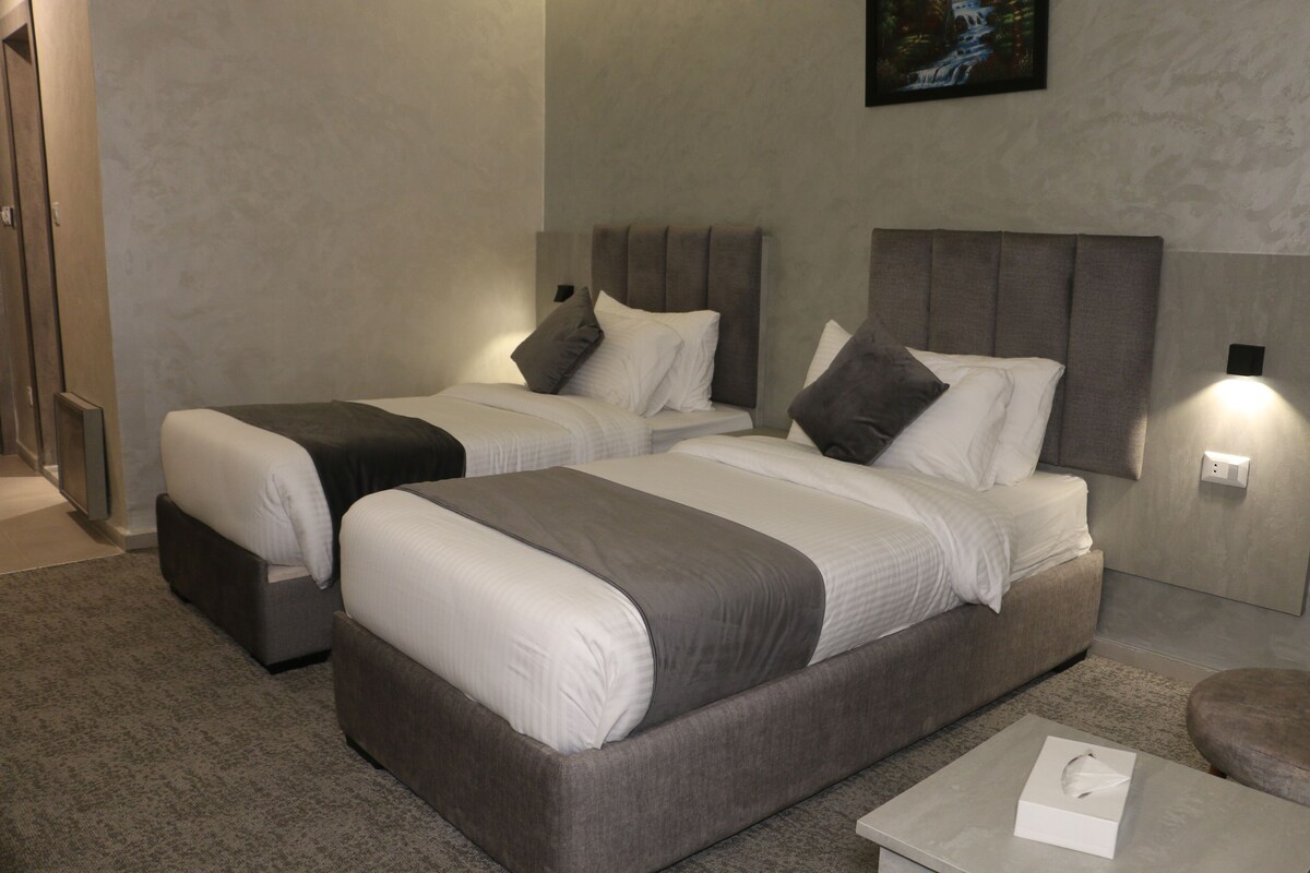 A hotel room with two beds, a small table, and a wall-mounted light.