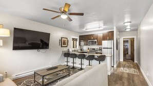 A modern living room with a flat-screen TV, a ceiling fan, and a kitchen area with a microwave and refrigerator.