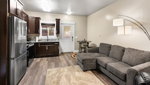 A modern kitchen with dark wooden cabinets, a stainless steel refrigerator, and a built-in oven. There is a gray sectional sofa and a small round table with chairs in the living area.