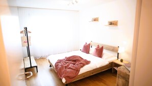 Una cama bien tendida con cabecera de madera, una mesita de noche con lámpara y un pequeño estante sobre la cama.