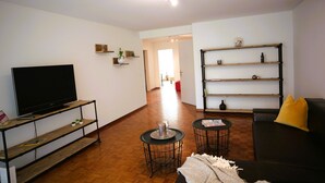 Una sala de estar con un televisor de pantalla plana sobre un soporte de madera, dos mesas redondas con sillas, un sofá negro con un cojín amarillo y una estantería con objetos decorativos.