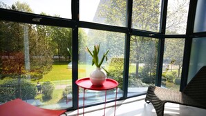 Una habitación moderna con ventanas amplias, una silla roja, un florero blanco con plantas y una mesa roja.