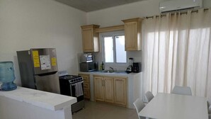 Eine Küche mit Kühlschrank, Herd, Mikrowelle und Wasserspender. Es gibt einen Esstisch mit Stühlen und ein Fenster mit Vorhängen.