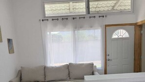 Ein heller, gut beleuchteter Raum mit einer Couch, einem Esstisch mit Stühlen und einem großen Fenster mit Vorhängen.