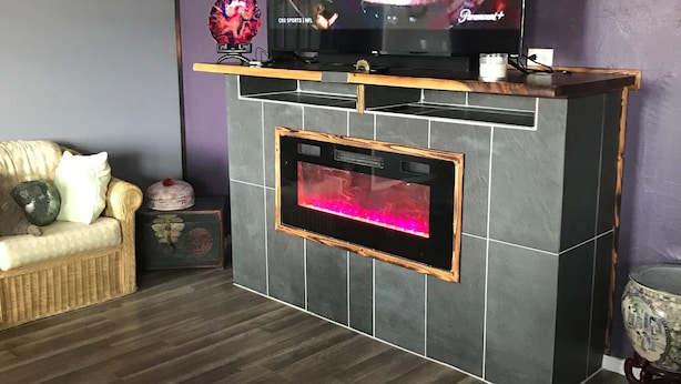 Un salon avec un téléviseur à écran plat fixé au-dessus d’une cheminée sombre, un canapé en osier et un vase décoratif posé sur une table d’appoint.