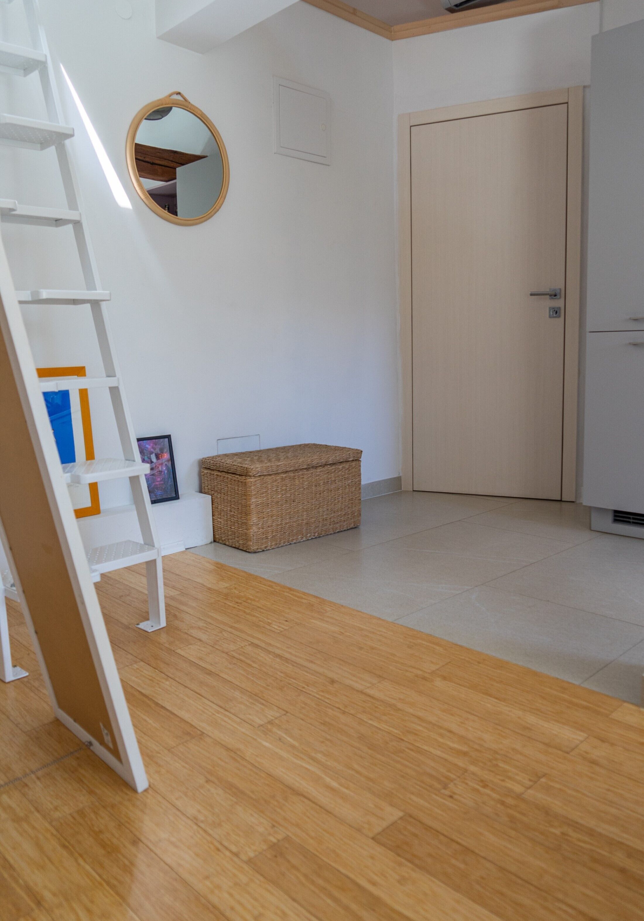 Una habitación con piso de madera, una puerta blanca, un armario blanco, una cesta de mimbre, un cuadro enmarco y una escalera blanca.