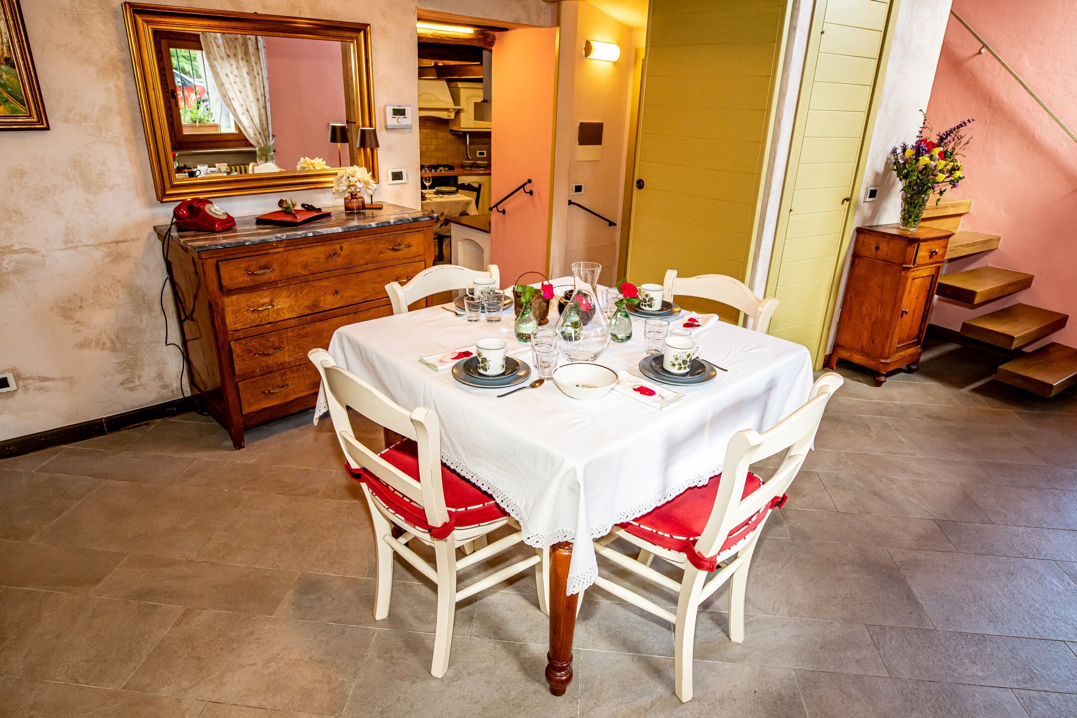 A dining area with a table set for four, white chairs with red cushions, a wooden cabinet, and a mirror on the wall.