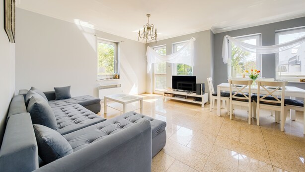 A modern living room with a sectional sofa, a TV, a dining table with chairs, and large windows with sheer curtains. - Apartamenty Sun & Snow Morskie Oko (Miedzyzdroje)