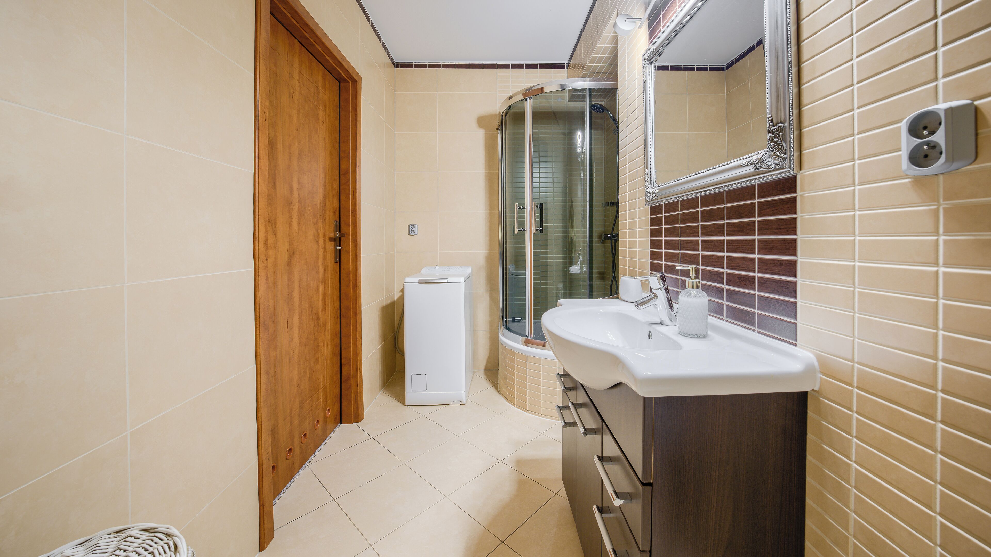 Une salle de bain avec une porte en bois, un espace de douche en verre, un lavabo blanc et un miroir doté d’un cadre décoratif.