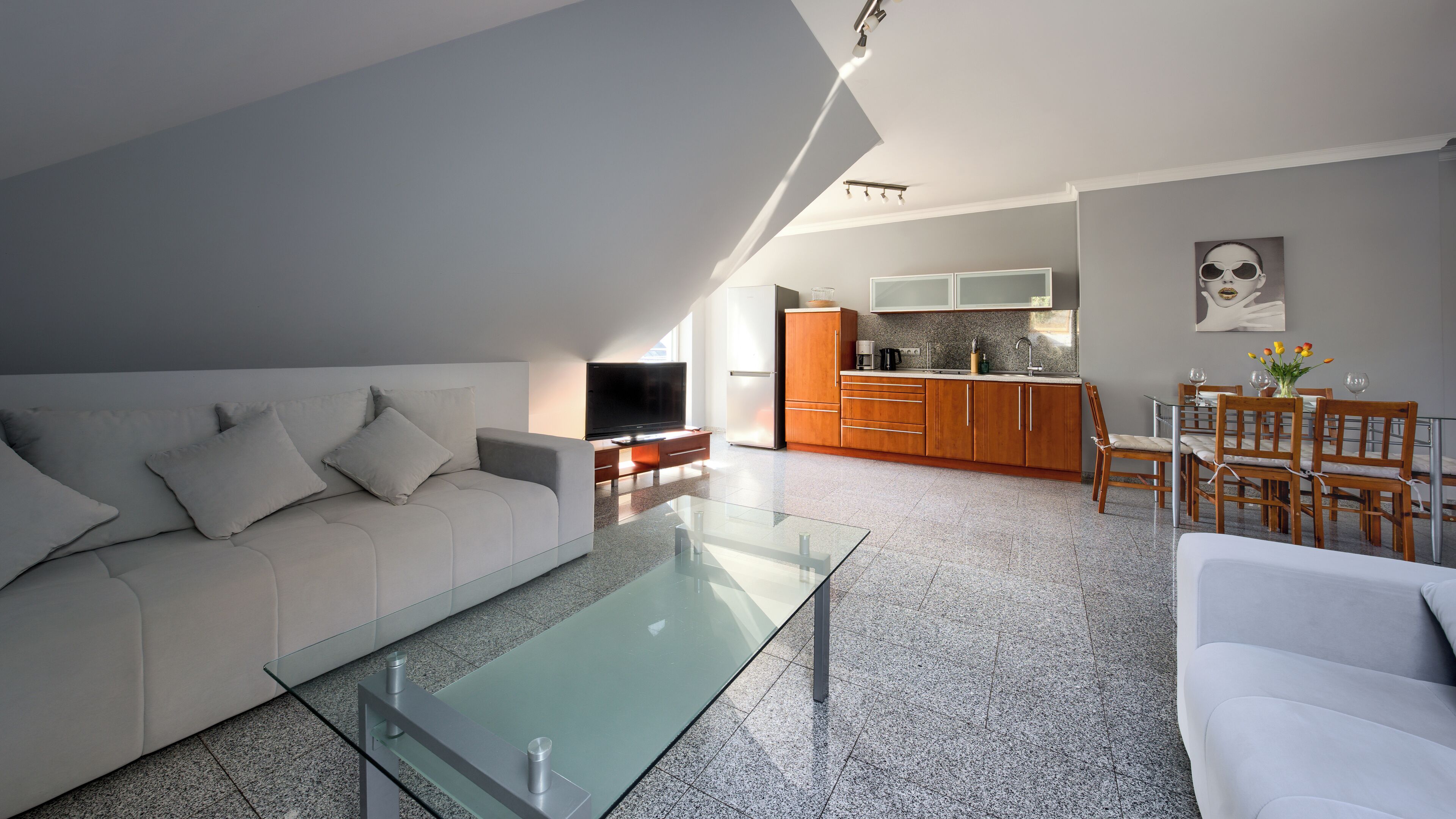 A modern living room with a glass coffee table, a white sofa, and a kitchen area with wooden cabinets and a dining table.