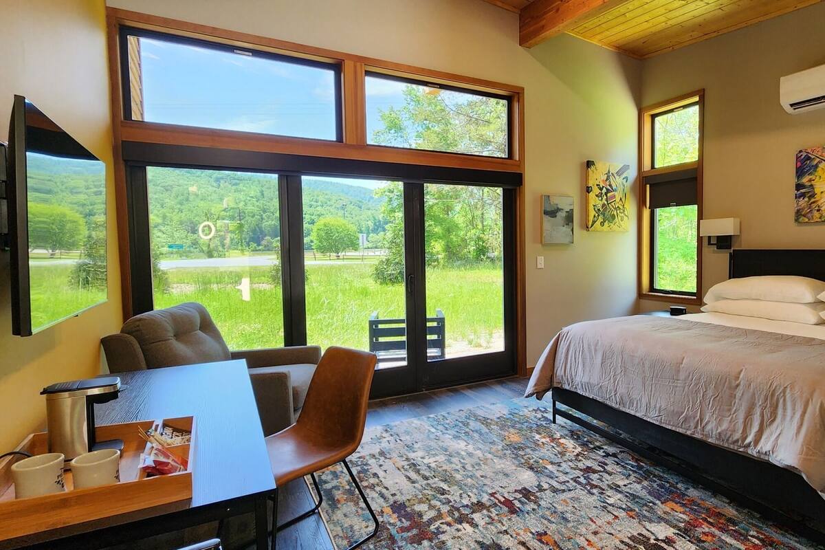 A bedroom with a bed, a desk with a chair, a large window with a view of greenery, and an air conditioning unit.