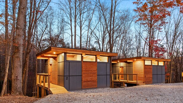Une cabane moderne, avec une façade en bois et une allée en gravier, entourée d’arbres.