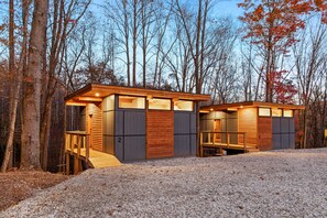Une cabane moderne, avec une façade en bois et une allée en gravier, entourée d’arbres.