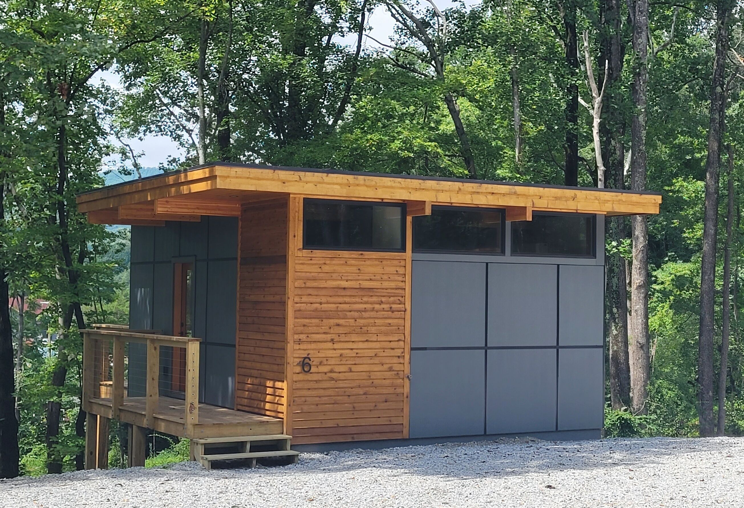 Een klein houten vakantiehuisje met een terras, grote ramen en een grijze deur, omringd door bomen.