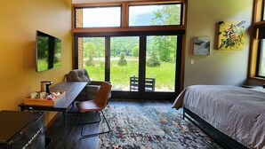 Une chambre avec un lit, un bureau et une chaise, une grande fenêtre donnant sur un paysage verdoyant, et un tableau accroché au mur.