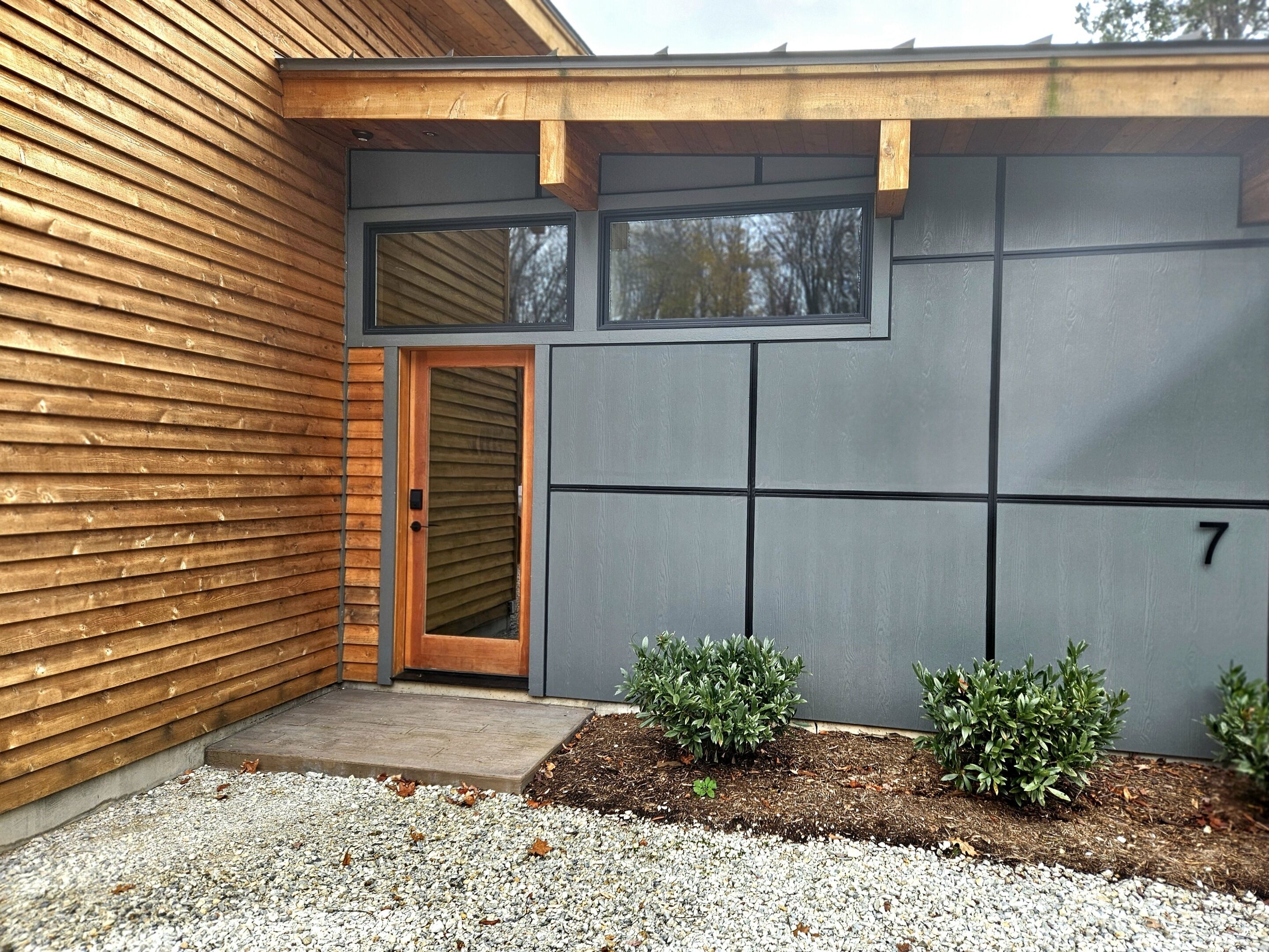 A modern building with a wooden exterior, a grey wall with a number '7', and a small garden area with gravel and plants.