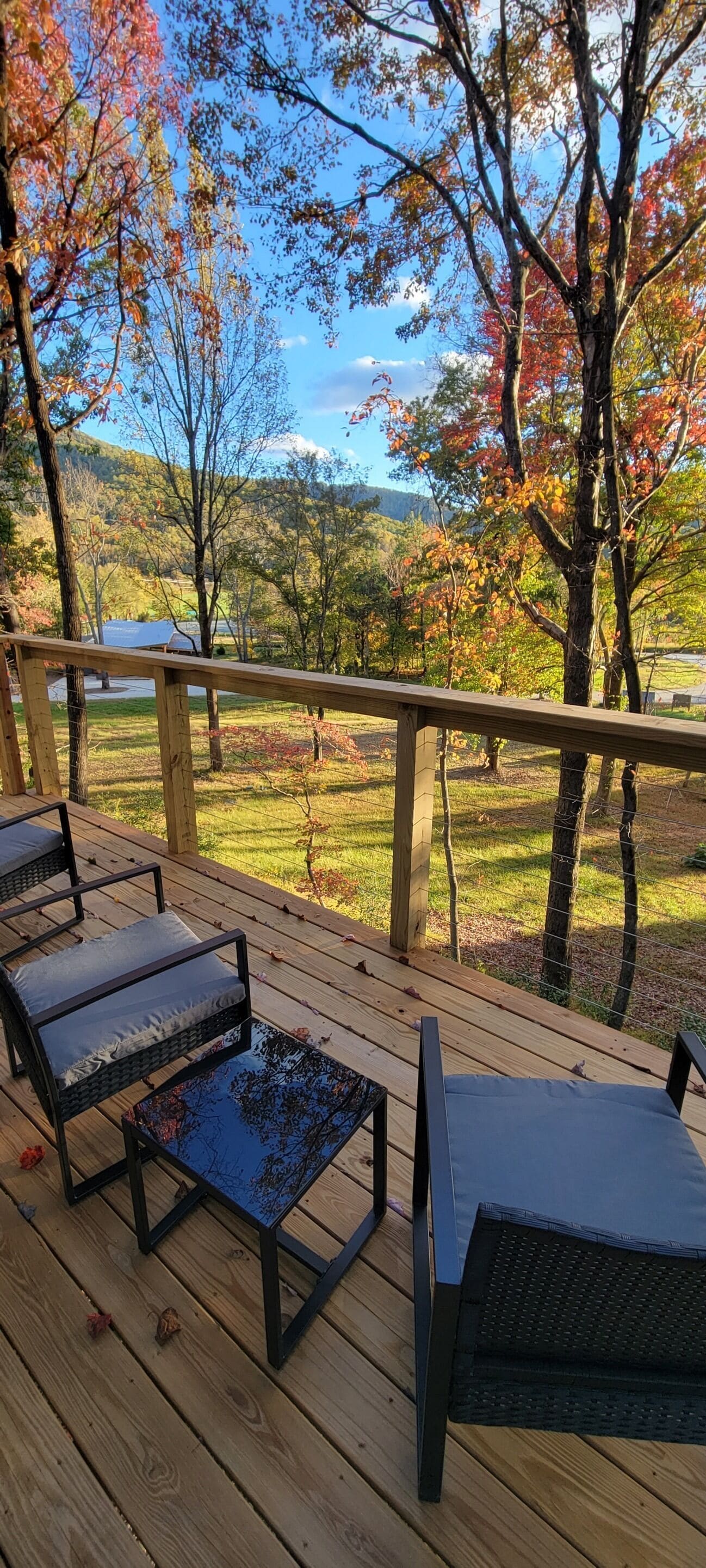 A wooden deck with four chairs and a table, overlooking a scenic landscape with autumn-colored trees.
