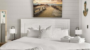 Une chambre à coucher avec un lit blanc, des oreillers blancs, une tête de lit blanche et un tableau représentant une scène de plage au mur.