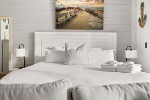 A bedroom with a white bed, white pillows, a white headboard, and a beach scene painting on the wall.