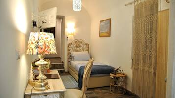 A small, cozy bedroom with a bed, a desk with a lamp, a chair, and a decorative wall hanging.