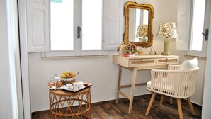 Una habitación con tocador de madera, un espejo, una silla de mimbre y una mesita con una bandeja de pasteles.