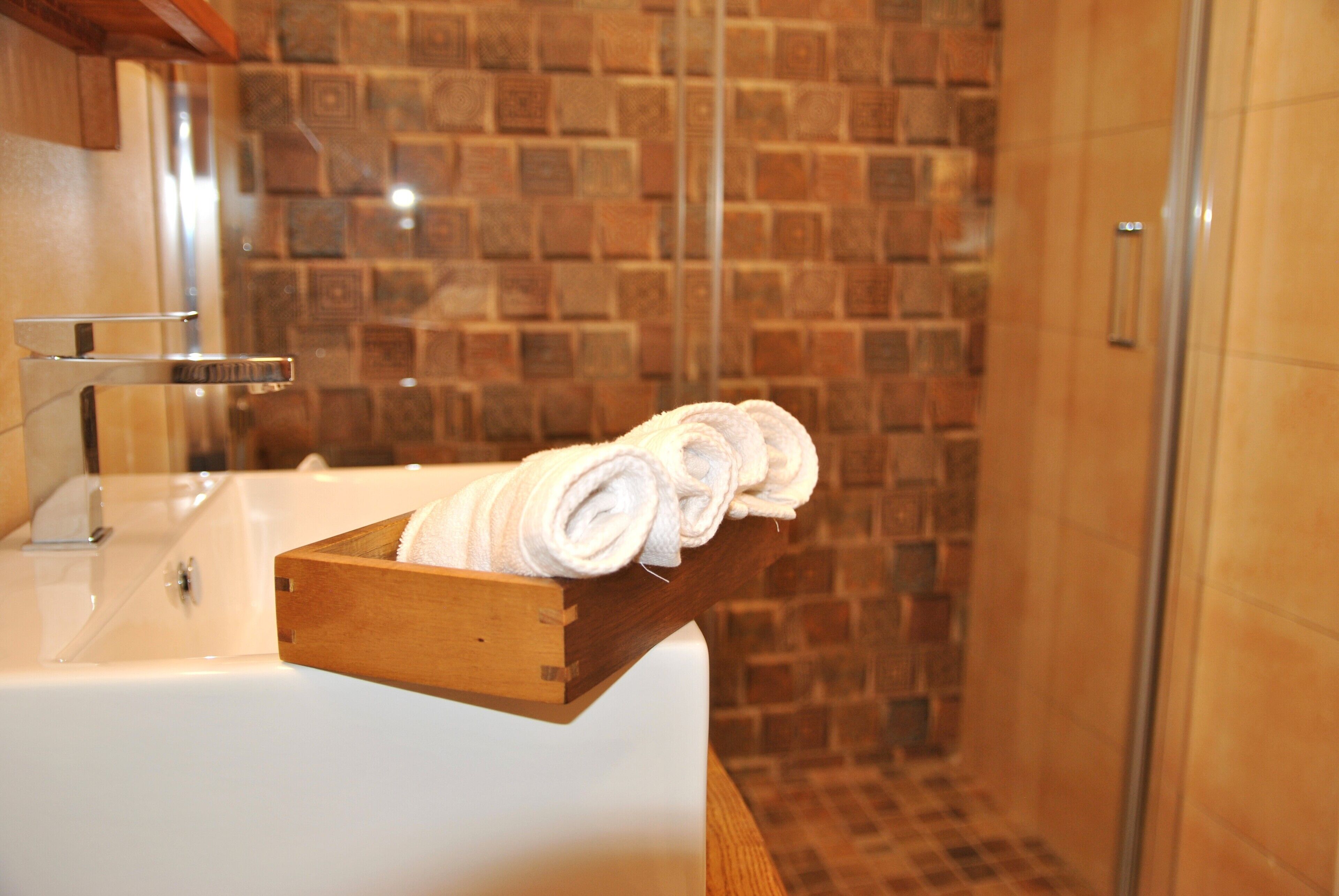 Un baño con un perchero de madera sosteniendo toallas enrolladas, un lavamanos blanco y una zona de ducha con pared de azulejos.