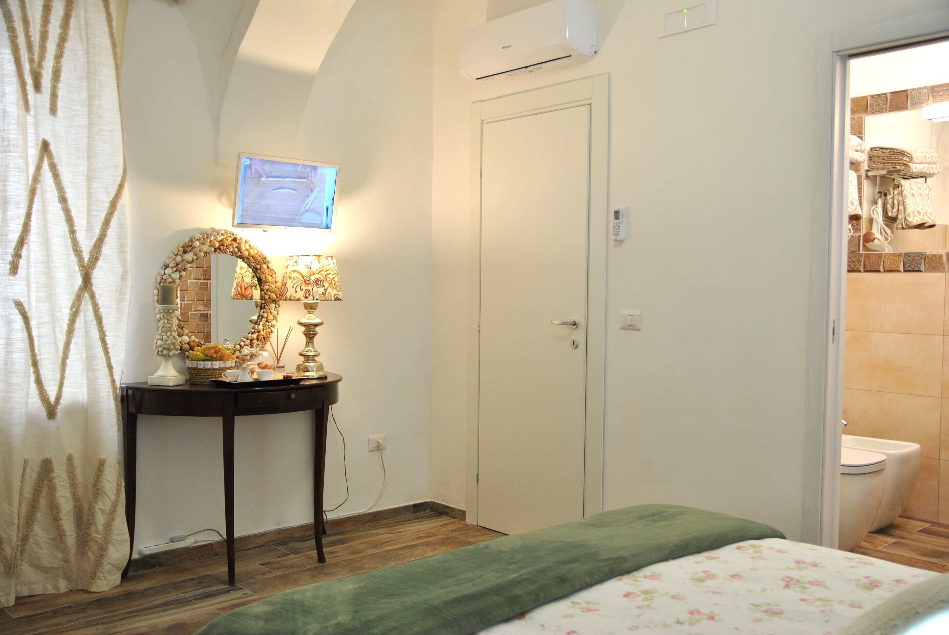 A bedroom with a bed, a bedside table with a lamp, a mirror, and a door to the bathroom.