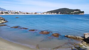 Una playa costera con agua azul cristalina, costa rocosa y una colina distante con edificios.