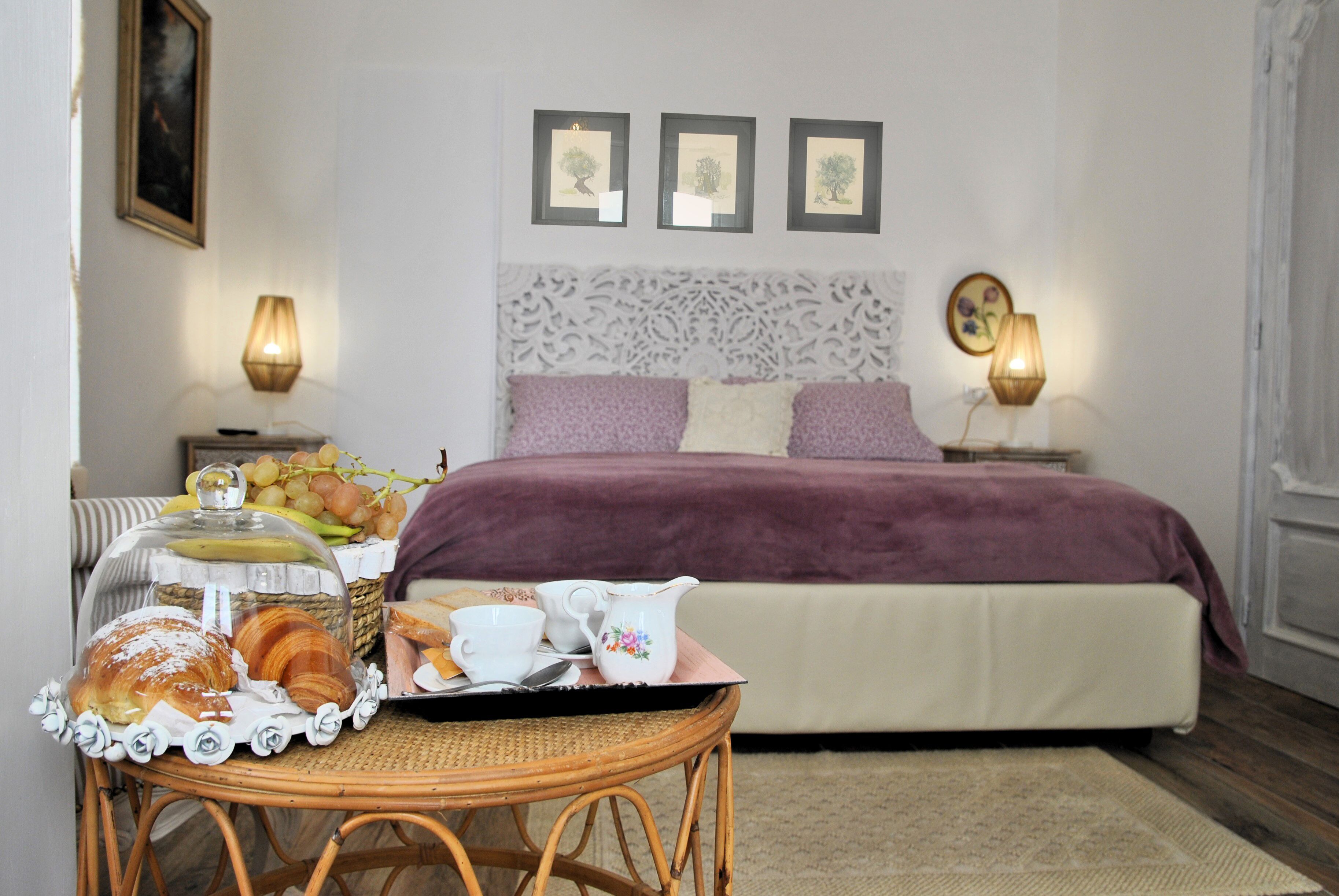 Een netjes opgemaakte slaapkamer met een bed, nachtlampjes, een decoratief hoofdeinde en een ontbijtkarretje met gebak en een koffiepot.