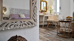 Una habitación con un espejo grande y ornamentado, una silla de mimbre, una pequeña mesa redonda con una cesta y un escritorio con una lámpara.