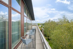 Ein Balkon mit Metallstühlen und einem Glasgeländer, der auf einen Garten und Gebäude blickt.