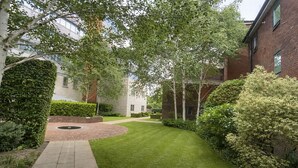 A well-maintained courtyard with a paved walkway, neatly trimmed grass, and mature trees providing shade. - Roomspace -The Courtyard Penthouse (London)