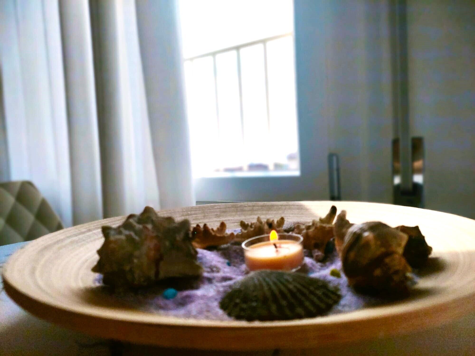 A candle on a plate with decorative items in a room with a window and curtains.