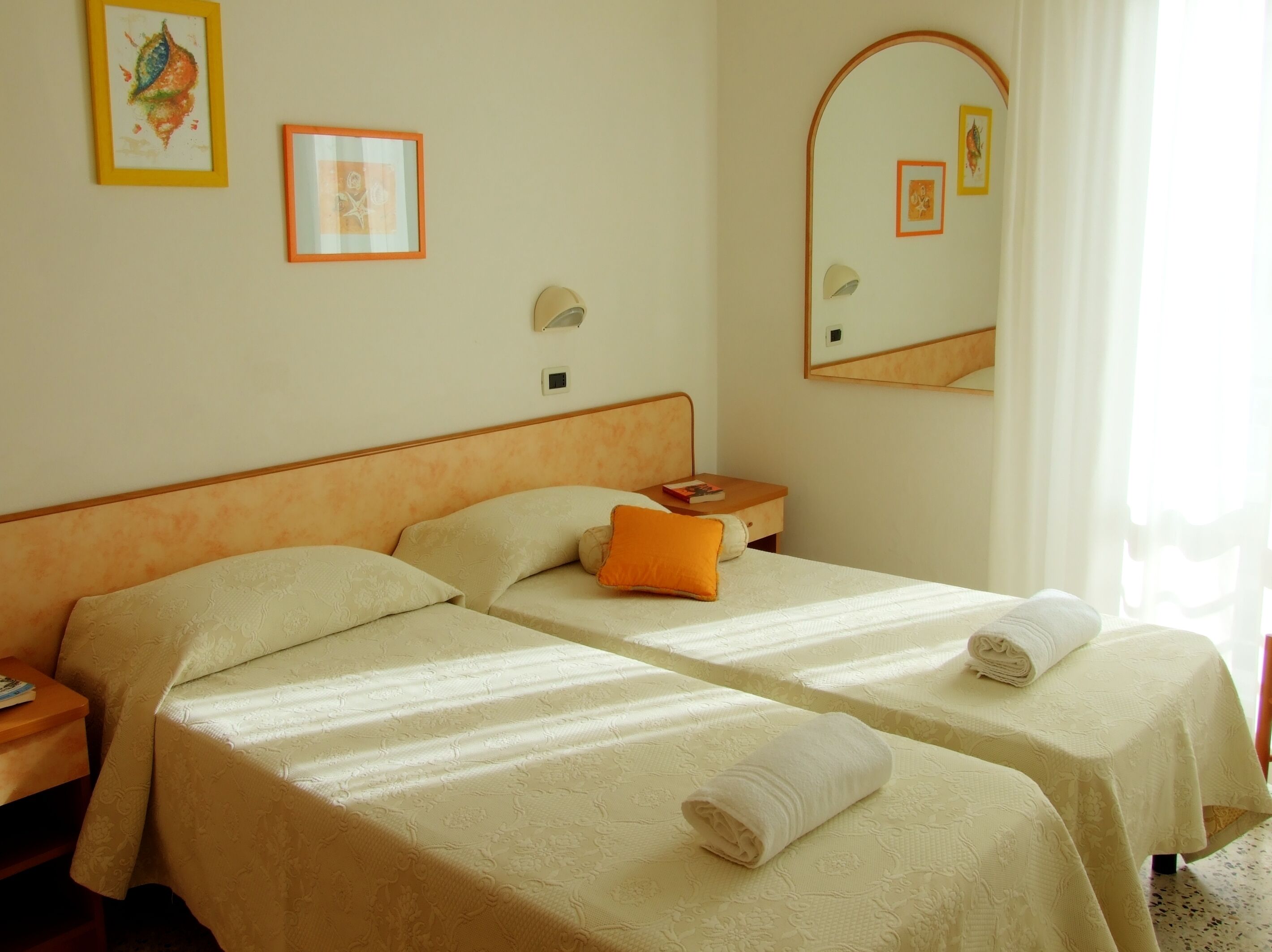 A hotel room with two beds, each with a pillow and a rolled towel. There is a nightstand with a book and a lamp. The room has a mirror and wall art.