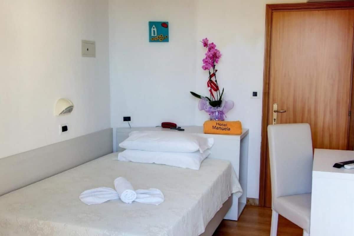 Hotellihuone, jossa on sänky, tuoli, pieni pöytä, kyltti, jossa lukee ”Hotel Manuel”, ja seinälle asennettu valaisin.