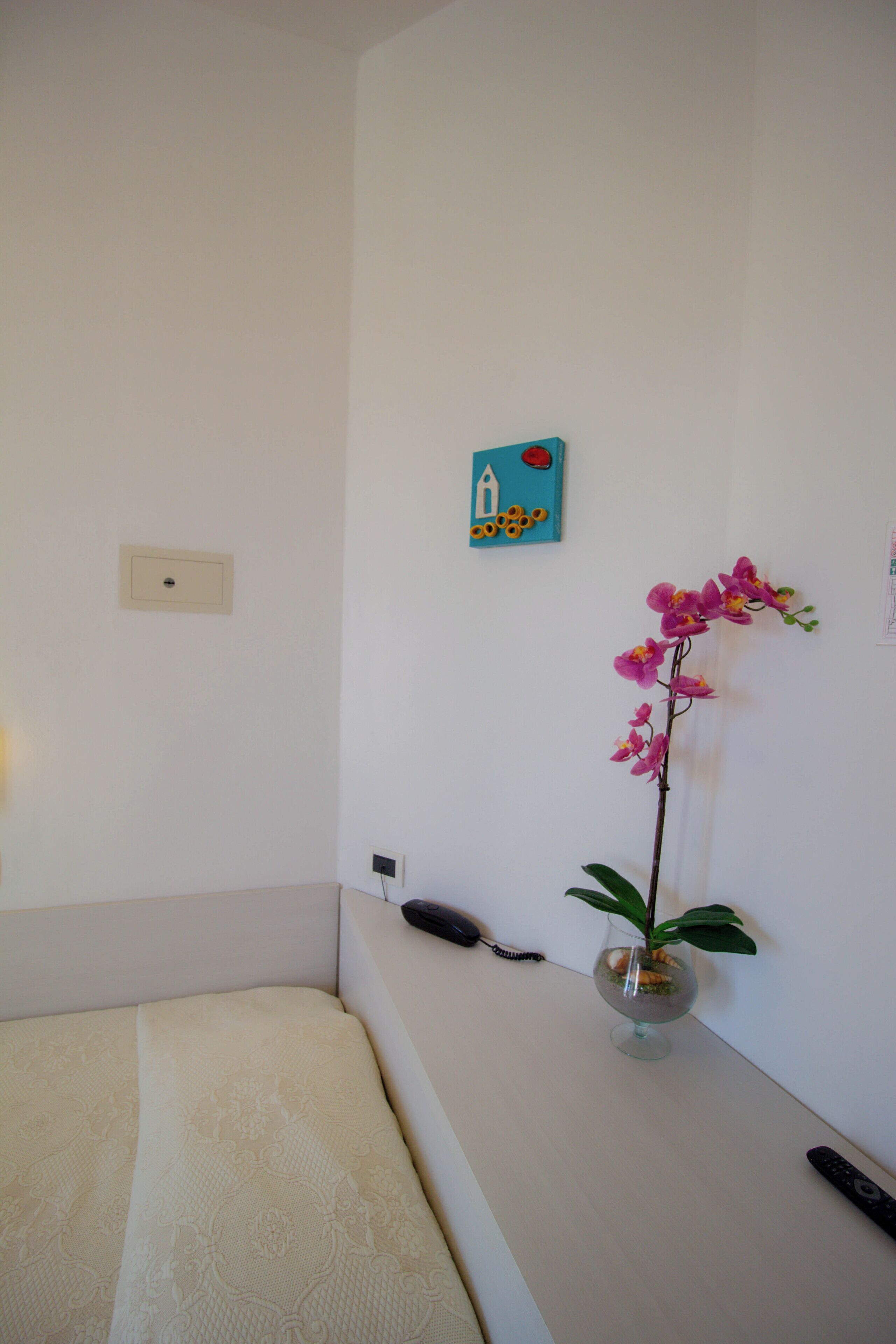 A room with a bed, a vase with an orchid, a remote control, and a wall-mounted phone.