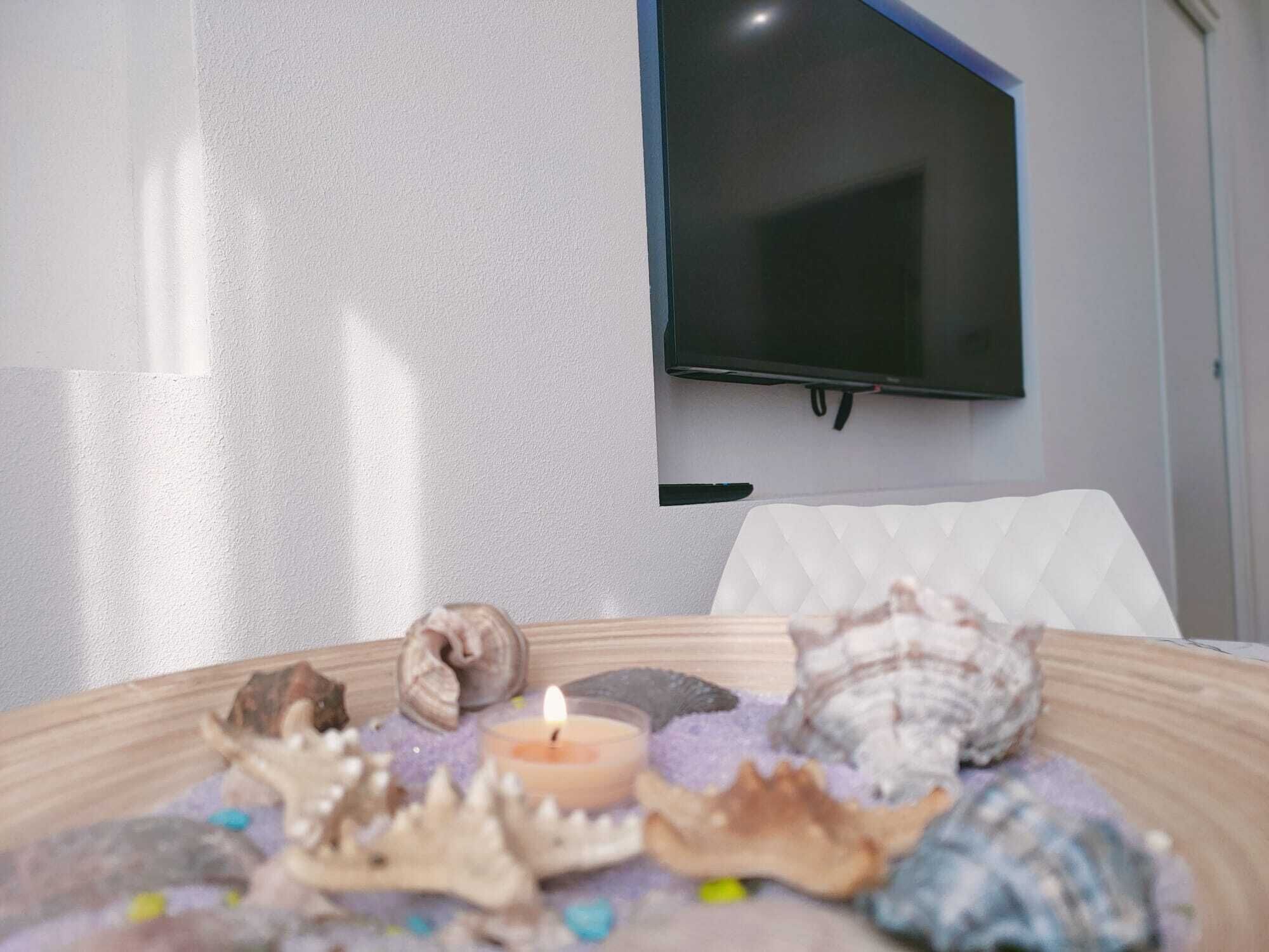 A room with a flat-screen TV mounted on the wall, a white cushioned chair, and a table with seashells and a lit candle.