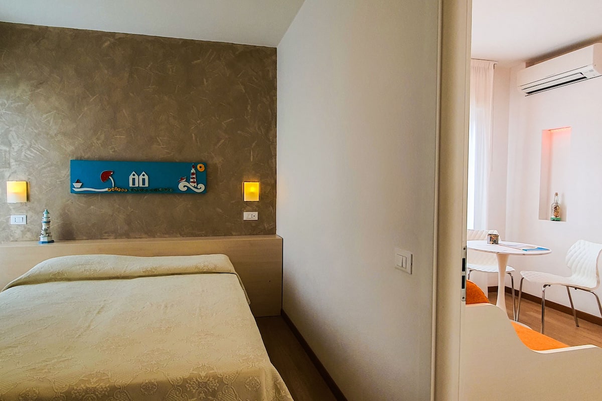 Hotellihuone, jossa on sänky, seinälle kiinnitetty taideteos, pieni pöytä tuoleineen ja ilmastointilaite.