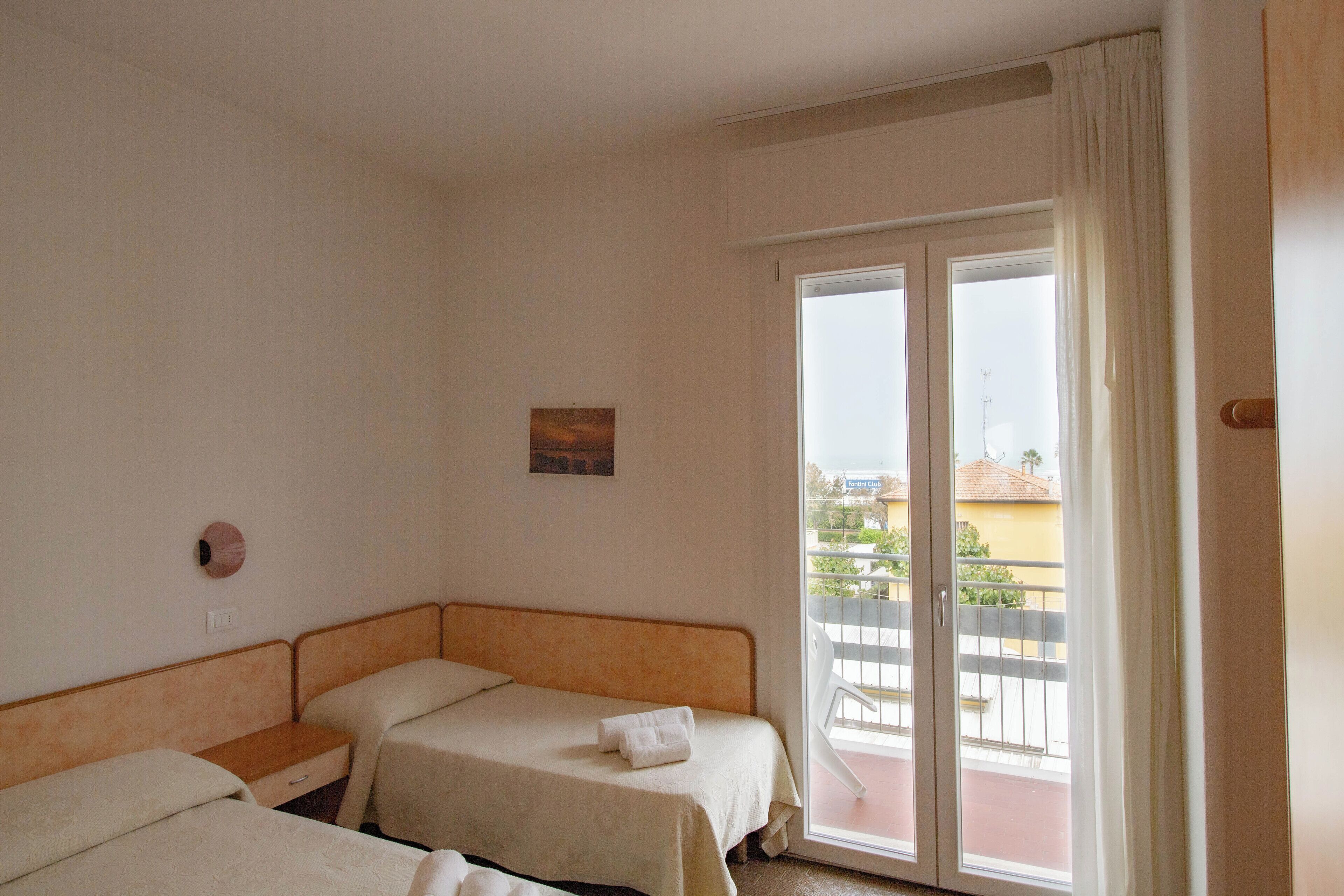 Habitación de hotel con dos camas individuales, una mesita, un reloj de pared y una ventana con vistas a un balcón y edificios.
