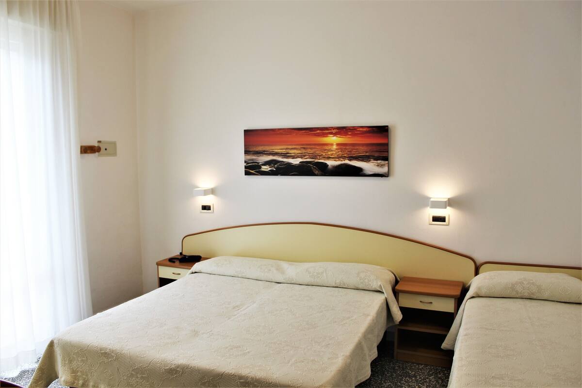 Hotellihuone, jossa on kaksi sänkyä, yöpöytä, lamppu ja seinälle ripustettu kuva auringonlaskusta meren yllä.