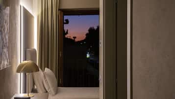 Ein Hotelzimmer mit Bett, Schreibtisch mit Lampe und Blick auf den Abendhimmel durch eine geöffnete Tür.