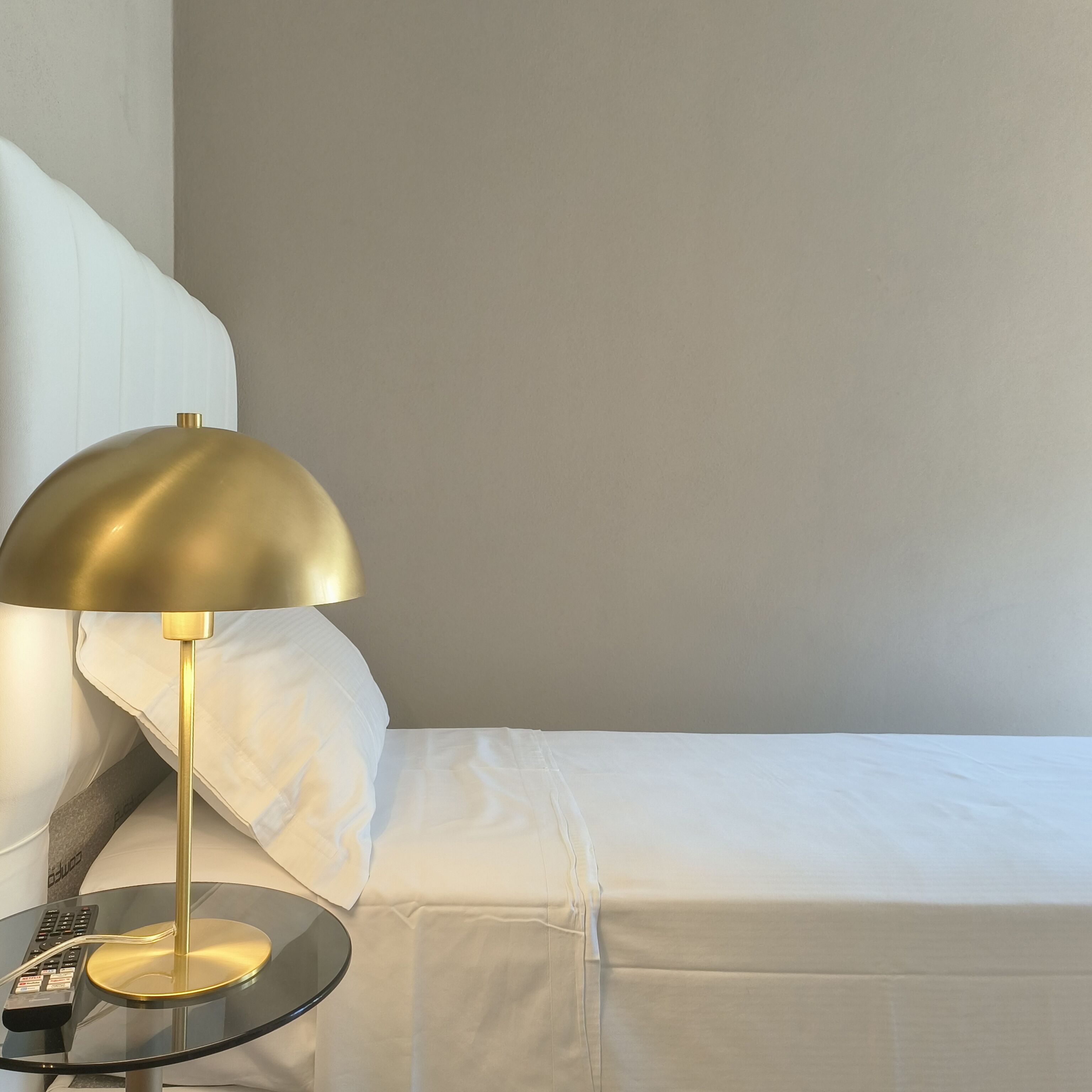 Una cama bien tendida con sábanas blancas, una lámpara dorada sobre una mesa negra y un control remoto en la mesa.