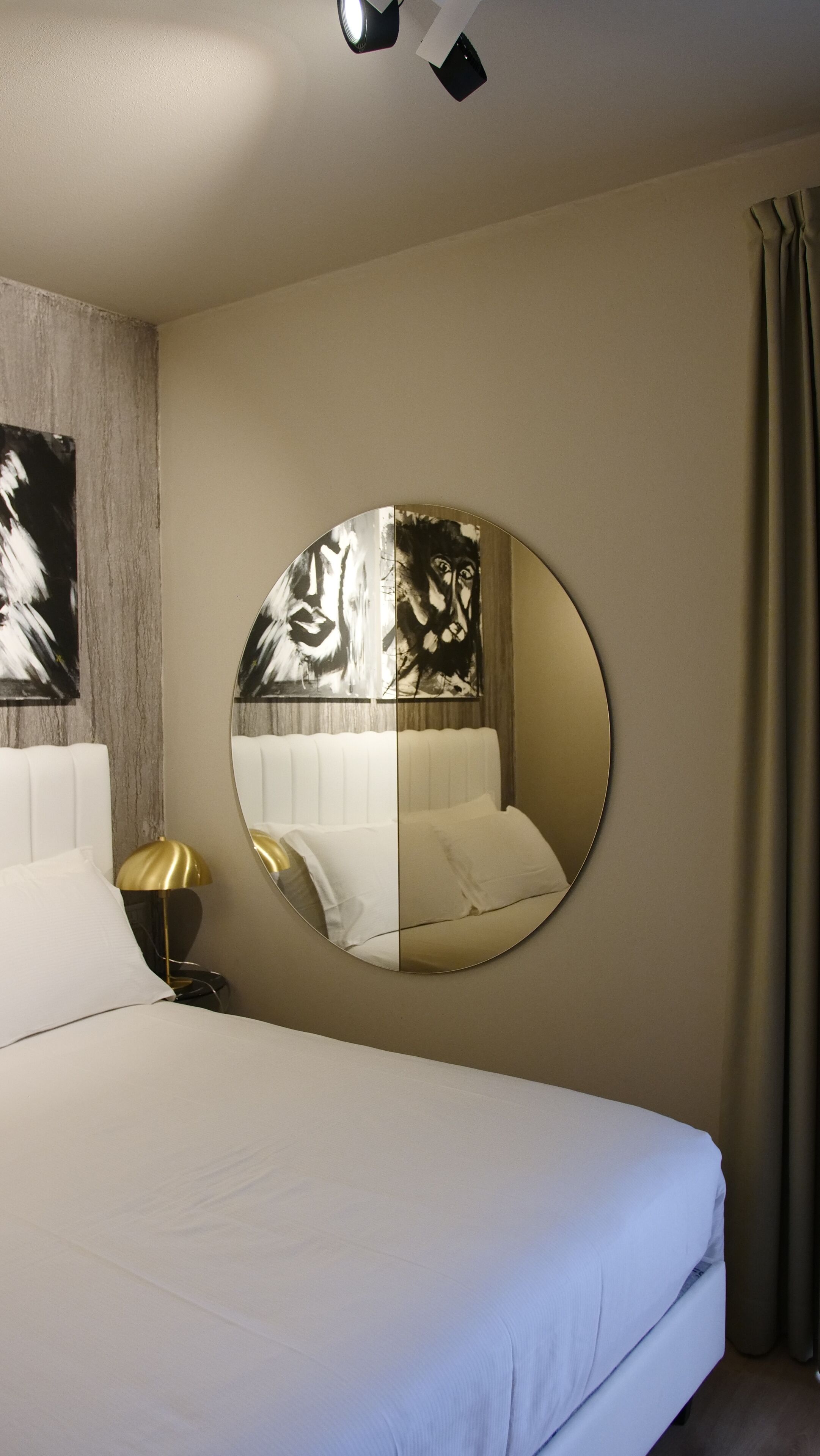 A hotel room with a large round mirror, a bed with white linens, a nightstand with a lamp, and a painting on the wall.