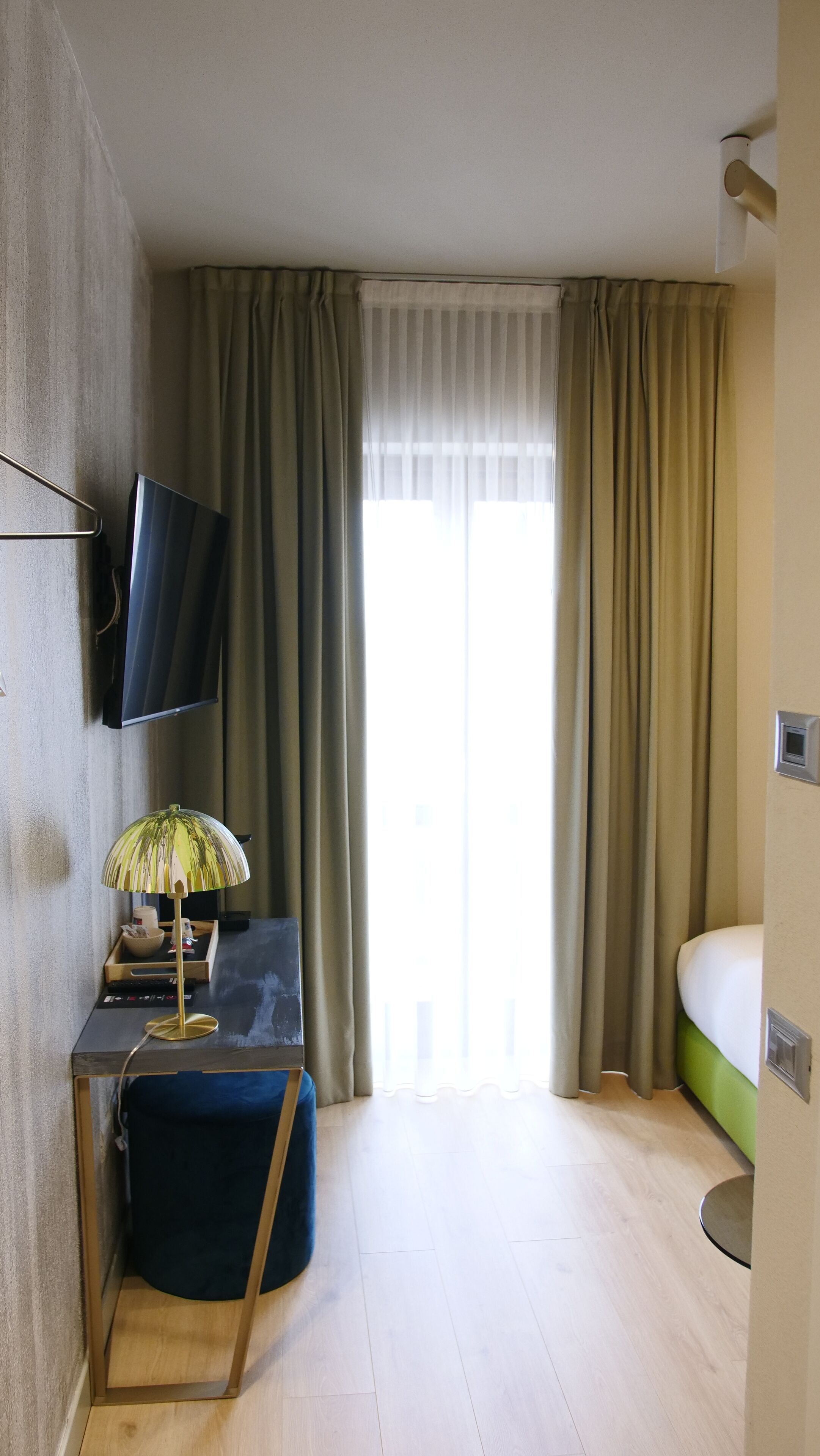 Habitación de hotel con cama, escritorio con lámpara, televisor montado en la pared y ventana con cortinas.