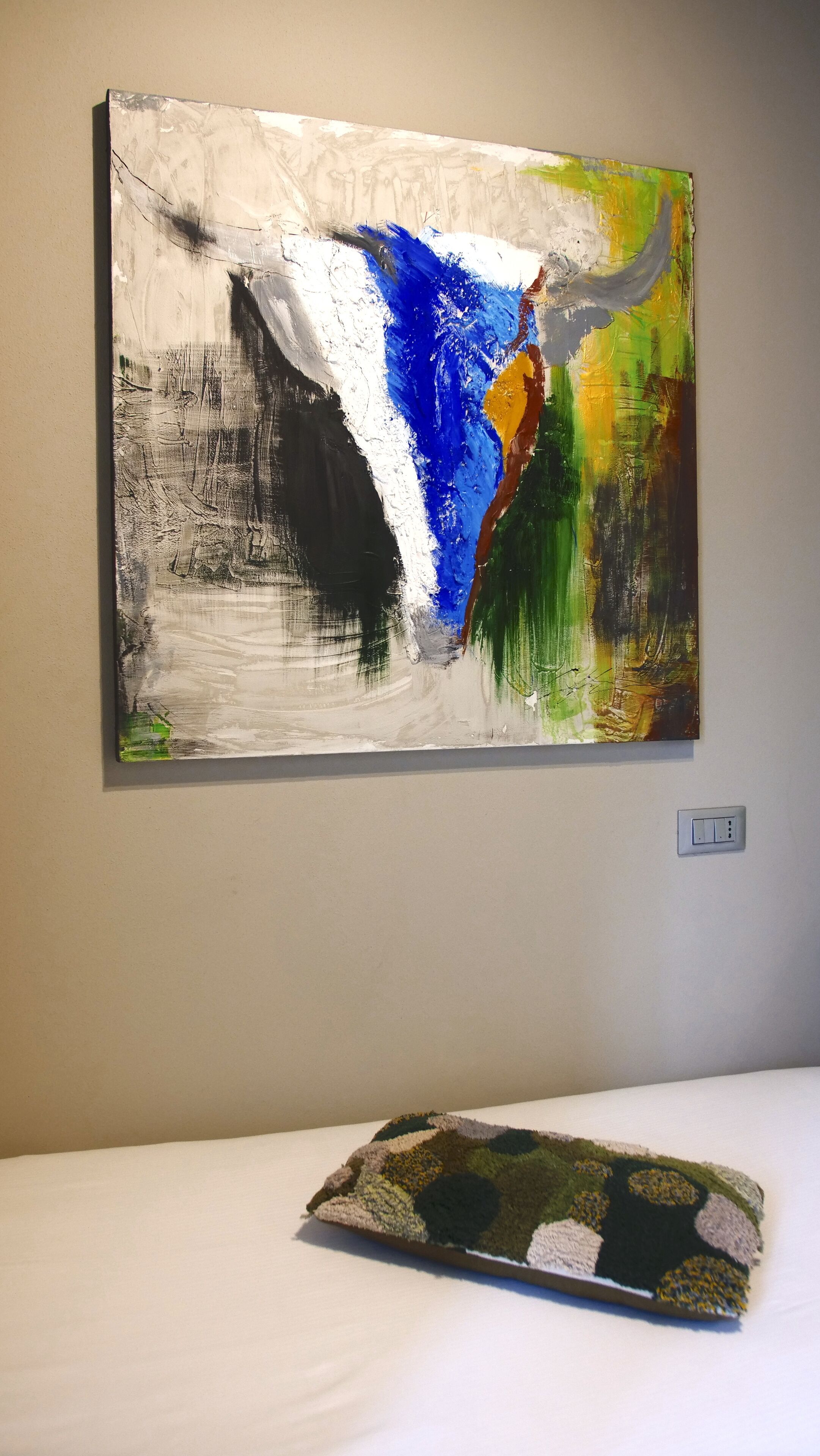 A room with a large abstract painting on the wall, a white bed, and a textured pillow.