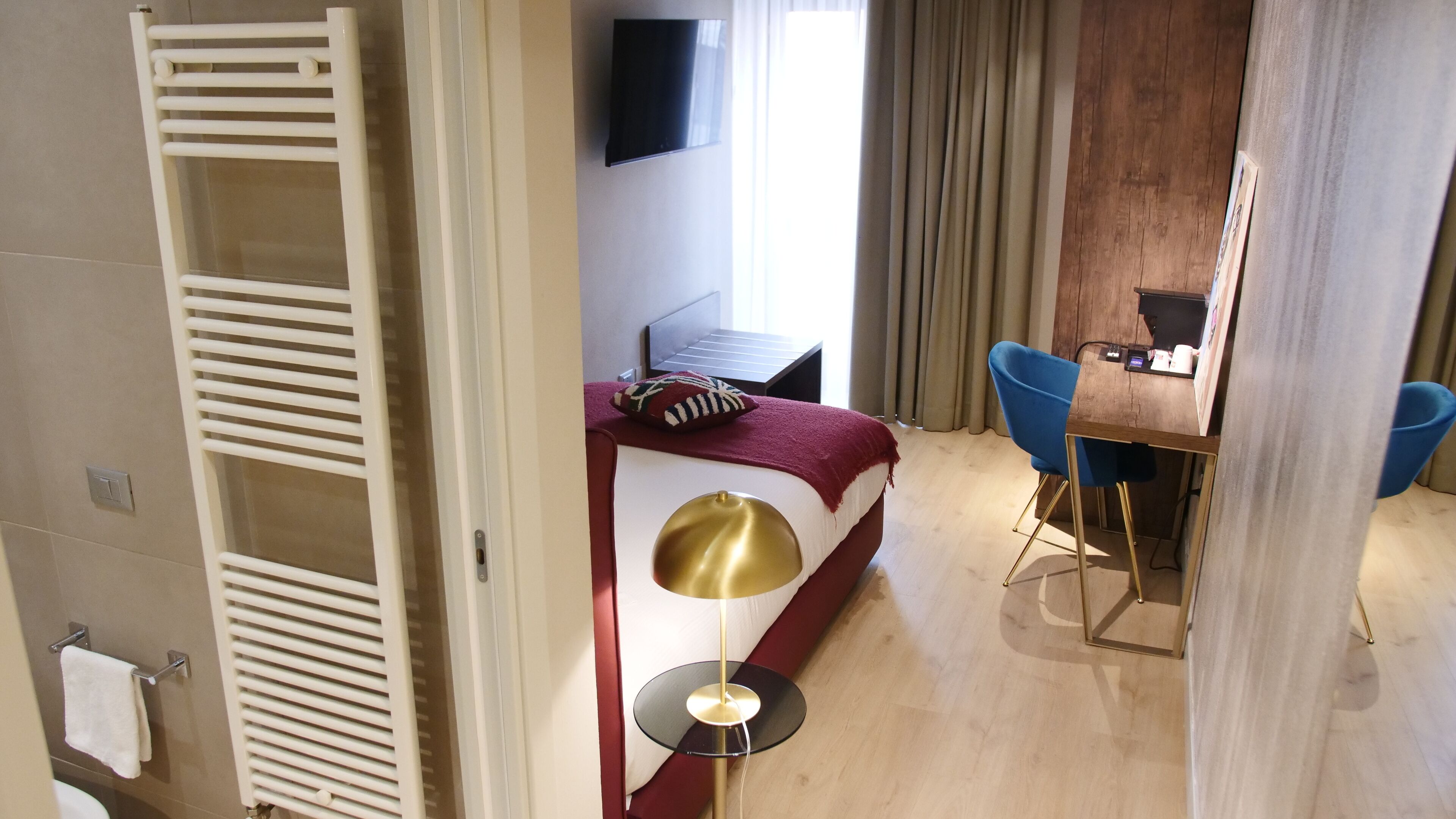 Camera d'albergo con un letto, una scrivania, una sedia, una televisione e un bagno con appendi-asciugamani.