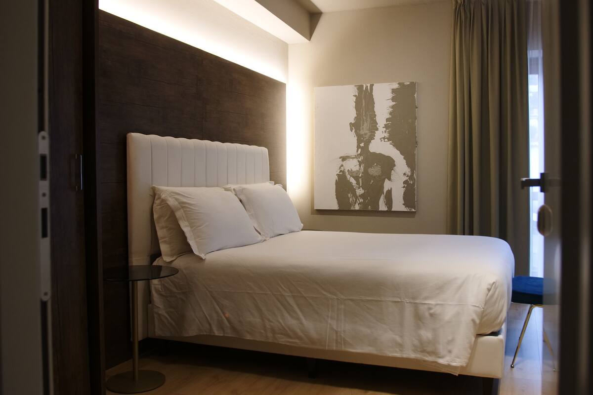 Une chambre moderne avec un grand lit, une tête de lit, une table de chevet et une œuvre d’art accrochée au mur.