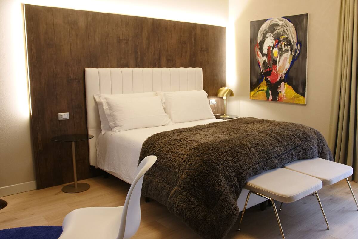 Une chambre à coucher moderne avec un grand lit, une table de chevet avec une lampe, une chaise et un tableau au mur.